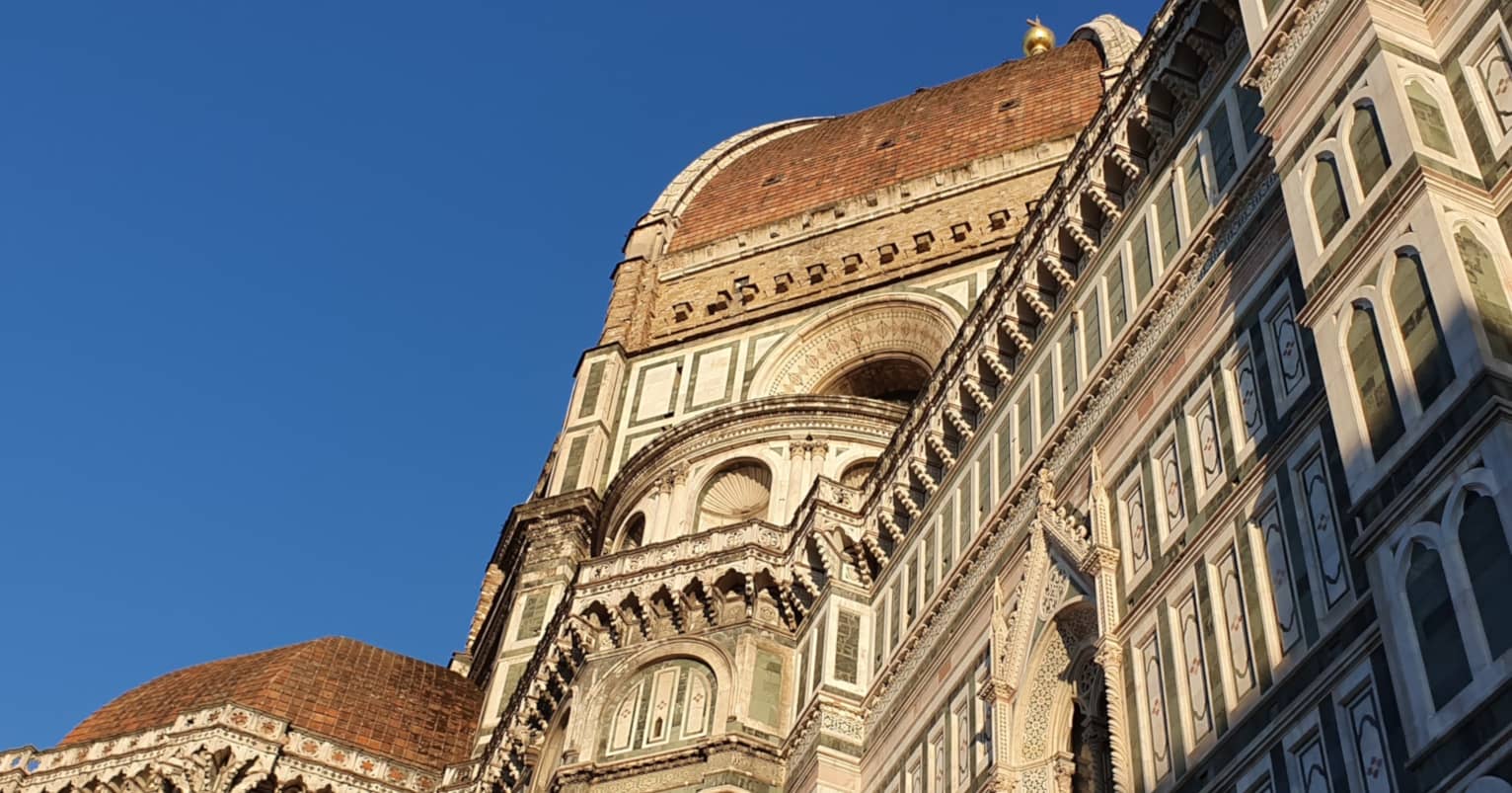 Contre-plongée sur le Duomo de Florence : la coupole de brique de Brunelleschi et la façade de marbre vert, blanc et rose, dans une lumière rasante de fin de journée, sur un ciel sans nuage.